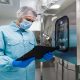 caucasian scientist blue lab uniform stand near big steel machine with control panel make notes about machine (1) (1)