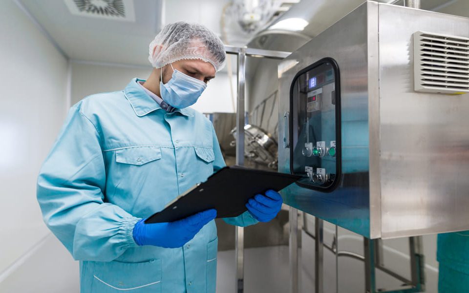 caucasian scientist blue lab uniform stand near big steel machine with control panel make notes about machine (1) (1)